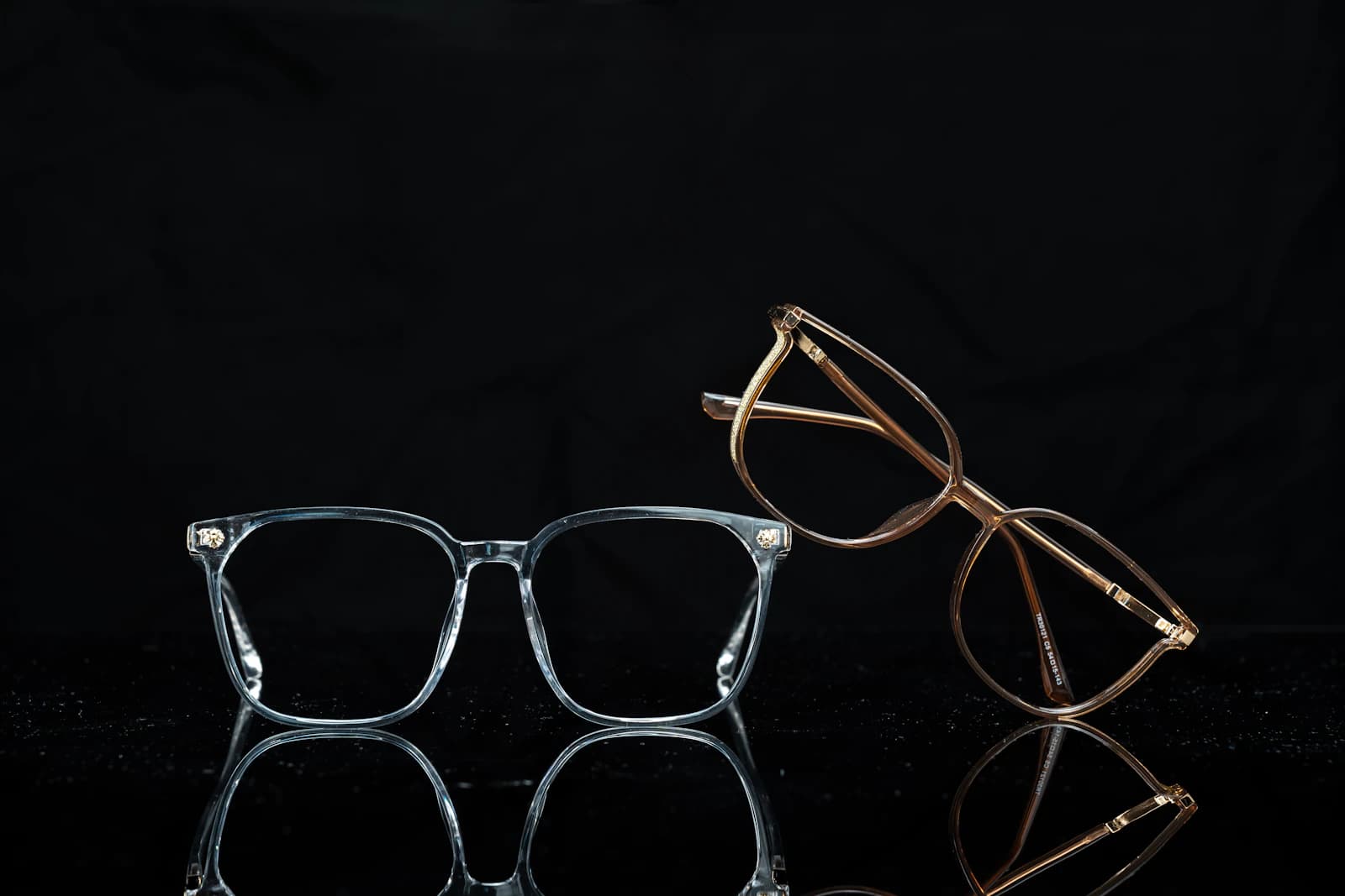 Two acetate optical frames with small decorative metal accents at the hinges, set on a black reflective surface